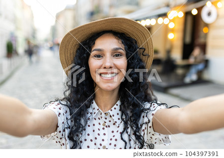 Young beautiful Latin American woman walking in evening city, woman looking into smartphone camera, using video call app, talking with friends smiling. Young beautiful Latin American woman walking in evening city, woman looking into smartphone camera, using video call app, talking with friends smiling. 104397391
