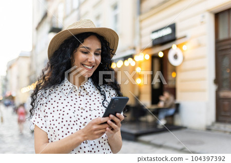 A beautiful young woman walks through the evening city in a hat, a smiling Latin American woman holds a smartphone in her hands. A tourist with curly hair types a message and browses online pages on A beautiful young woman walks through the evening city in a hat, a smiling Latin American woman holds a smartphone in her hands. A tourist with curly hair types a message and browses online pages on 104397392