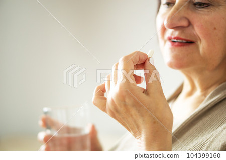 Close up of a retired woman in casual clothes at home holding pill and glass water.  104399160