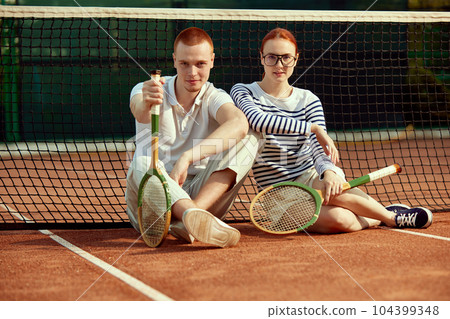 Handsome young man and beautiful woman in casual, stylish clothes posing with tennis racket on court on daytime Handsome young man and beautiful woman in casual, stylish clothes posing with tennis racket on court on daytime 104399348