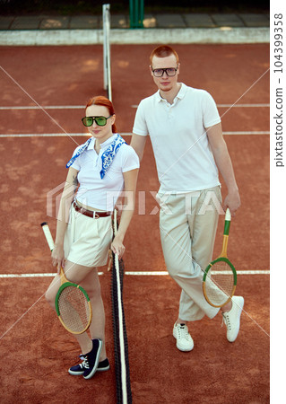 Full-length top view image of beautiful young woman and handsome man in casual, stylish clothes posing on tennis court on daytime Full-length top view image of beautiful young woman and handsome man in casual, stylish clothes posing on tennis court on daytime 104399358