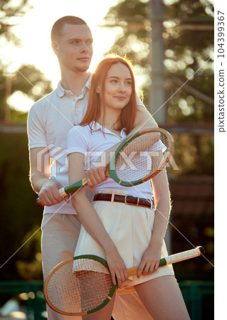 Beautiful young redhead man and woman in stylish, casual, light clothes posing with tennis racket on warm day during sunset time 104399367