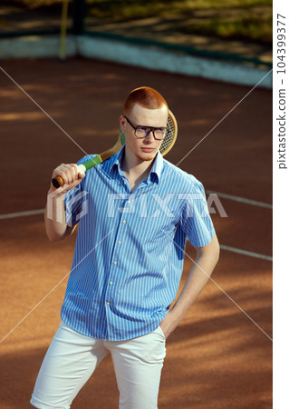 Portrait of handsome redhead young man in stylish casual clothes and glasses posing on opendoor tennis court with tennis racket Portrait of handsome redhead young man in stylish casual clothes and glasses posing on opendoor tennis court with tennis racket 104399377