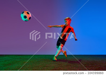 One sportive child, boy in red uniform playing, training football over dark blue background in neon. Concept of action, sportive lifestyle, team game, health, energy 104399860