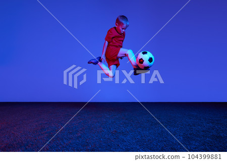 Male beginner soccer player in sports uniform in action, motion with ball over dark blue background in neon. Concept of sport, health, action, ad Male beginner soccer player in sports uniform in action, motion with ball over dark blue background in neon. Concept of sport, health, action, ad 104399881