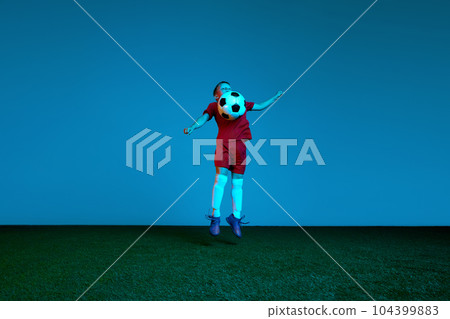 Studio shot of little boy, soccer player in sports uniform jumping with football ball over dark blue background in neon. Concept of sport, health, action, ad Studio shot of little boy, soccer player in sports uniform jumping with football ball over dark blue background in neon. Concept of sport, health, action, ad 104399883