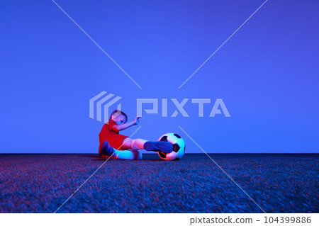 Male beginner soccer player in sports uniform in action, motion with ball over dark blue background in neon. Concept of sport, health, action, ad 104399886