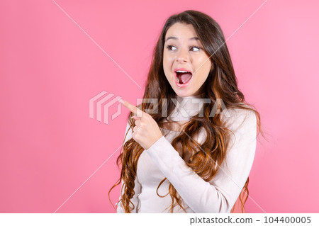 Portrait of a young lady stands on a pink backdrop, glancing left and gesturing towards a copy space 104400005