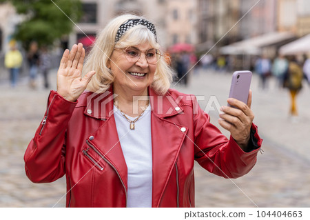 Senior woman blogger taking selfie on smartphone video call online with subscribers in city street 104404663