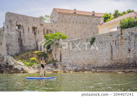 Young women Having Fun Stand Up Paddling in blue water sea in Montenegro. Against the backdrop of the Old Town of Budva. SUP Young women Having Fun Stand Up Paddling in blue water sea in Montenegro. Against the backdrop of the Old Town of Budva. SUP 104407241