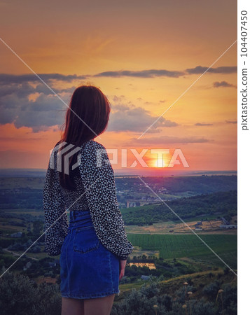 Rear view of a girl watching the sunset from the top over the valley. Natural summer evening scene Rear view of a girl watching the sunset from the top over the valley. Natural summer evening scene 104407450