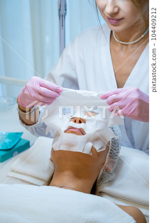 Beautician applying sheet mask on face of young woman client. 104409212