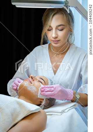 Blonde beautician doctor cleaning woman face with cotton pads, skin care procedures, rejuvenation. 104409242