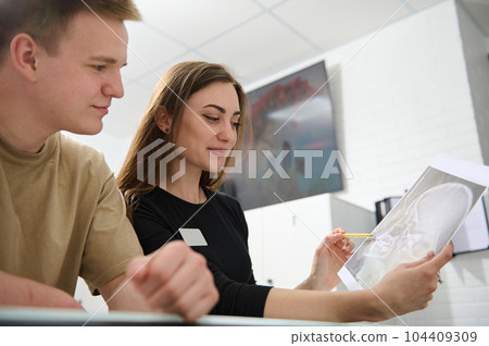 Cute female dentist doctor with Xray image and male patient standing at reception desk and discussing teeth treatment that needs to perform, scheduling the next appointment, in modern dentistry clinic 104409309