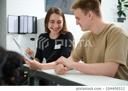Receptionist point of view: Smiling contented male patient and doctor dentist filling out forms, discussing dental treatment needs to perform, standing at the reception counter in a dentistry clinic 104409312
