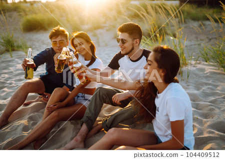 Young people sitting together at beach, drinking beer. Group of friends cheers with beers at the beach. Young people sitting together at beach, drinking beer. Group of friends cheers with beers at the beach. 104409512