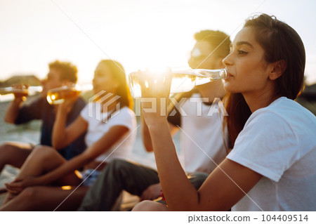 Young people sitting together at beach, drinking beer. Group of friends cheers with beers at the beach. 104409514