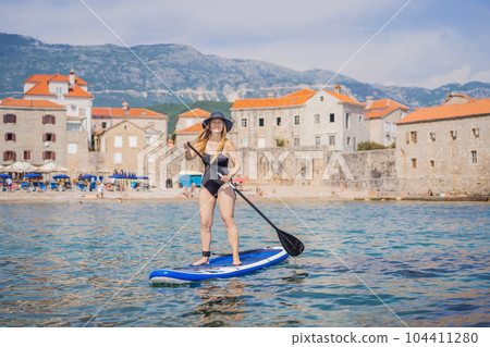 Young women Having Fun Stand Up Paddling in blue water sea in Montenegro. Against the backdrop of the Old Town of Budva. SUP 104411280