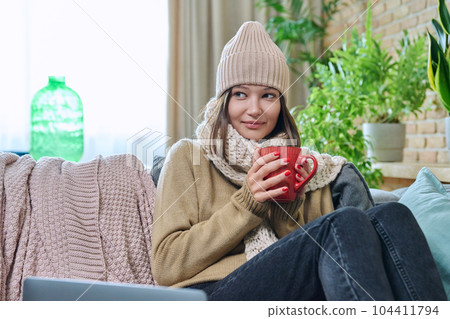 Young woman in sweater hat sitting at home, basking with cup of hot drink Young woman in sweater hat sitting at home, basking with cup of hot drink 104411794