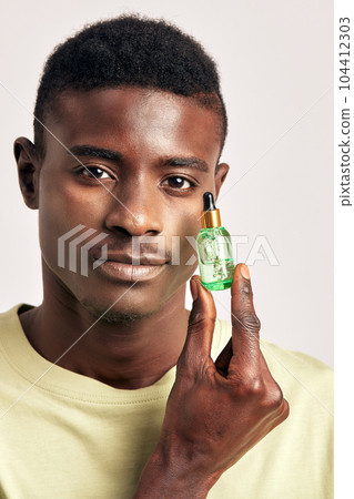 Young handsome confident African guy with a small vial of facial moisturizing gel in a studio. A black man caring for the skin to maintain his youthful and healthy appearance. Men's self-care. Young handsome confident African guy with a small vial of facial moisturizing gel in a studio. A black man caring for the skin to maintain his youthful and healthy appearance. Men's self-care. 104412303