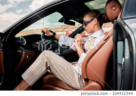 A young attractive business lady dressed in a white shirt and a beige vest is driving her luxury car. A confident blonde in sunglasses is driving to work or business negotiations and drinking smoothie A young attractive business lady dressed in a white shirt and a beige vest is driving her luxury car. A confident blonde in sunglasses is driving to work or business negotiations and drinking smoothie 104412344