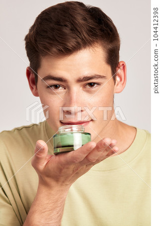 Young guy holding a vial of facial mask cream in his hands before applying it on his face during his skincare routine. Man caring for the skin to maintain his youthful and healthy appearance. Young guy holding a vial of facial mask cream in his hands before applying it on his face during his skincare routine. Man caring for the skin to maintain his youthful and healthy appearance. 104412398