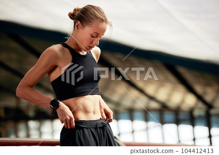 Young athlete girl is warming up in the stadium. Attractive slender confident blonde fixing her black tracksuit before training. Active lifestyle, sports and fitness. Young athlete girl is warming up in the stadium. Attractive slender confident blonde fixing her black tracksuit before training. Active lifestyle, sports and fitness. 104412413