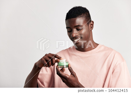 Young black guy holding a vial of facial mask cream in his hands before applying it on his face during his skincare routine. Man caring for the skin to maintain his youthful and healthy appearance. 104412415