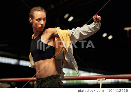 Young athlete girl getting ready for a workout at the stadium. Attractive slender blonde in black top taking off a yellow-gray sports jacket before training. Active lifestyle, sports and fitness. 104412416