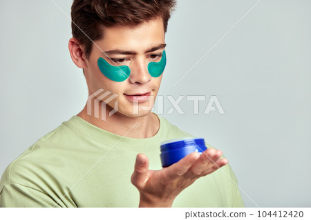 A millennial guy with brown hair practicing skincare with under eye hydrogel patches in a studio. Young smiling caucasian man applying cosmetic patches on his face. Men's self-care. 104412420