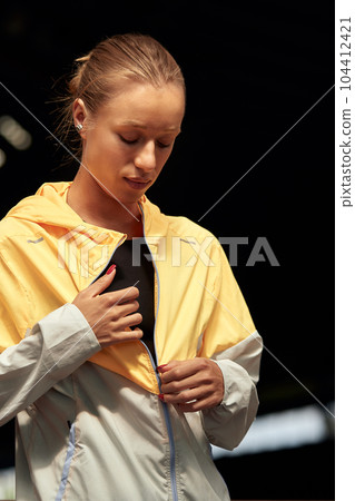Young athlete girl getting ready for a workout at the stadium. Attractive slender blonde taking off a yellow-gray sports jacket before training. Active lifestyle, sports and fitness. Young athlete girl getting ready for a workout at the stadium. Attractive slender blonde taking off a yellow-gray sports jacket before training. Active lifestyle, sports and fitness. 104412421