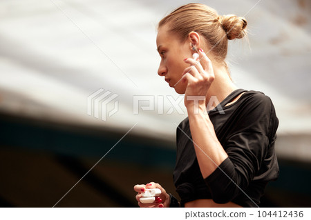 A cute young fitness girl takes out wireless headphones from a case and puts them in the ears. Attractive blonde in a black t-shirt listens to music and prepares for a workout. Active lifestyle. A cute young fitness girl takes out wireless headphones from a case and puts them in the ears. Attractive blonde in a black t-shirt listens to music and prepares for a workout. Active lifestyle. 104412436