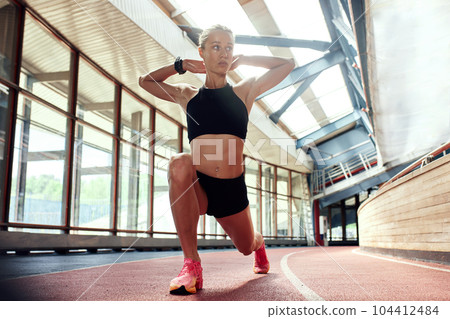 A young athlete girl is warming up before the race at the stadium. A slender blonde in a black tracksuit stretches her leg muscles before a competition on the track. Sports and recreation. 104412484