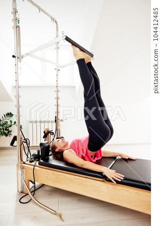 A young woman is doing Pilates on a reformer bed in a bright studio. A slender brunette in a pink top and black leggings does exercises to strengthen the muscles while lying on her back. 104412485