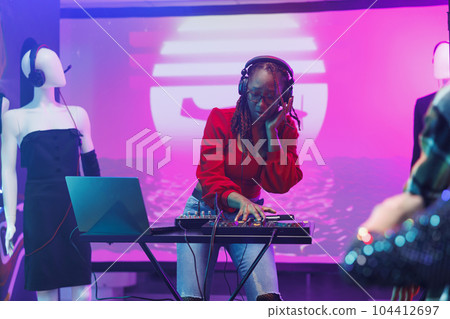 Woman dj in headphones mixing sound with controller while performing on stage with spotlights in nightclub. African american musician using digital music mixer during show in club 104412697