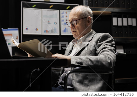 Experienced businessman with paraplegia in wheelchair checking bookkeeping analytical data paperwork. Old accountant executive in business company file cabinet repository filled with flowcharts and Experienced businessman with paraplegia in wheelchair checking bookkeeping analytical data paperwork. Old accountant executive in business company file cabinet repository filled with flowcharts and 104412736
