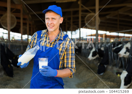 Smiling dairy farm owner pouring milk from bottle into glass in outdoor cowshed 104415269