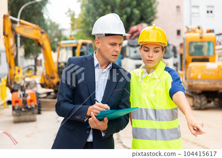 Young woman worker on a construction site shows something to a man engineer Young woman worker on a construction site shows something to a man engineer 104415617