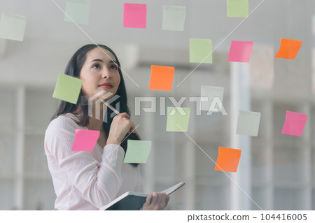 A professional millennial Asian businesswoman writing on colorful sticky notes and attaching them to a glass window in a creative office. Business planning and brainstorming strategy concept. 104416005