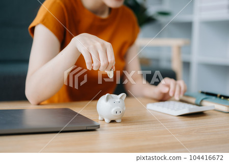 Asian women's hand saving a coin into piggy bank with wooden to calculateon and tablet on the table for business, finance, saving money and property investment concept.. 104416672