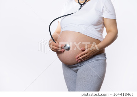 Close-up pregnant woman, expectant gravid mother holding hand on her belly, listening to heartbeat of her baby in womb with stethoscope, isolated on white background. Monitoring the fetus development Close-up pregnant woman, expectant gravid mother holding hand on her belly, listening to heartbeat of her baby in womb with stethoscope, isolated on white background. Monitoring the fetus development 104418465