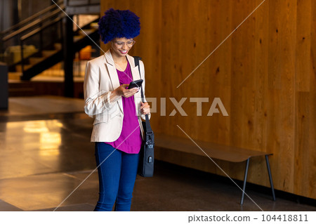 Happy biracial casual businesswoman with blue afro using smartphone in office corridor 104418811