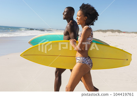 Happy, fit african american couple carrying surfboards walking on sunny beach to the sea 104419761
