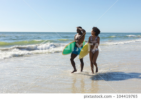 Happy, fit african american couple carrying surfboards walking on sunny beach by the sea, copy space Happy, fit african american couple carrying surfboards walking on sunny beach by the sea, copy space 104419765