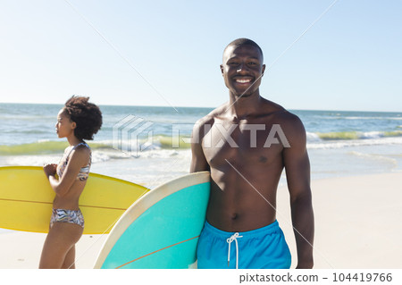 Portrait of happy african american man holding surfboard standing on sunny beach with female surfer Portrait of happy african american man holding surfboard standing on sunny beach with female surfer 104419766