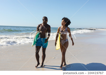 Happy, fit african american couple carrying surfboards walking on sunny beach by the sea 104419767