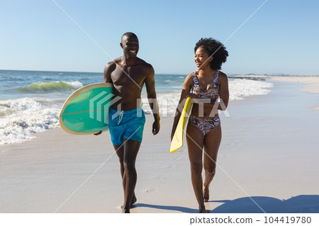 Happy, fit african american couple carrying surfboards walking on sunny beach by the sea 104419780