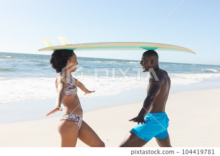 Happy african american couple balancing surfboard on heads, smiling on sunny beach 104419781