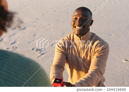 Happy african american man kneeling with ring and proposing to girlfriend on sunny beach 104419890