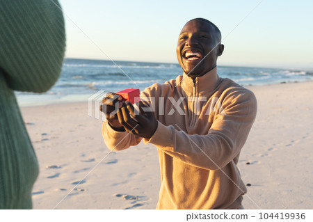 Happy african american man kneeling with ring and proposing to girlfriend on sunny beach 104419936